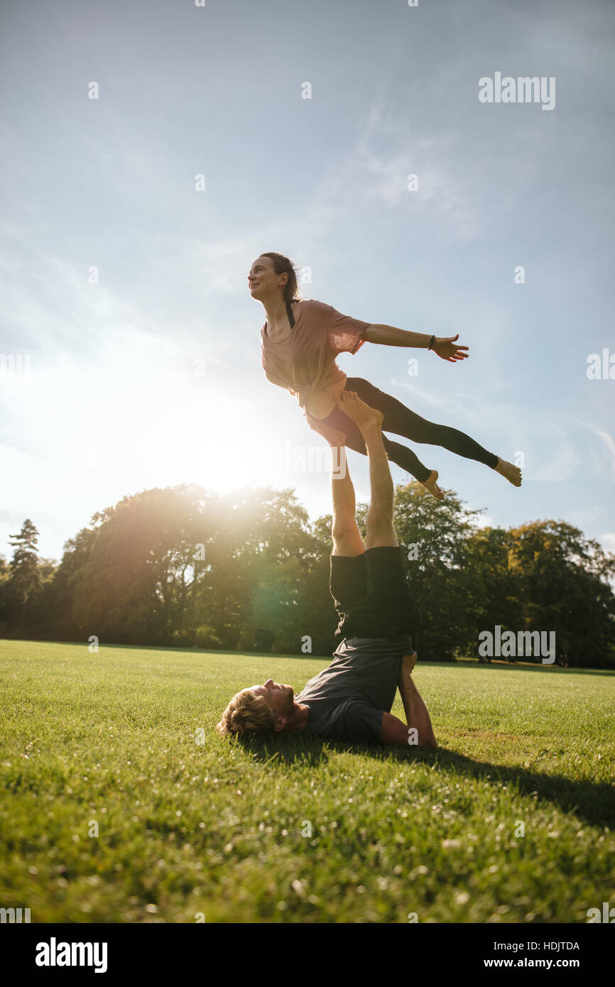 Disparo vertical de montar pareja joven haciendo yoga acrobático. Hombre tumbado sobre el césped y equilibrar la mujer en sus pies en el parque. Foto de stock