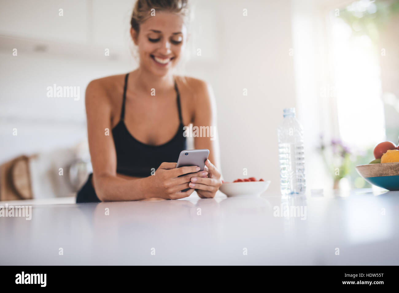 Hermosa mujer joven de pie en la encimera de la cocina y la lectura de mensajes de texto en su teléfono móvil. Foto de stock