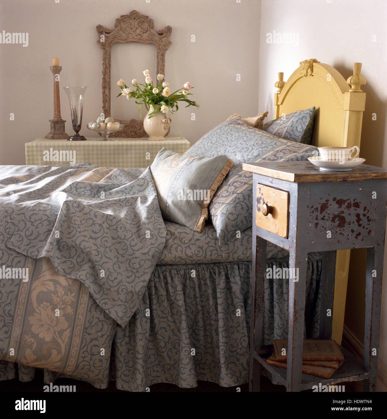 Pequeña pintada angustiado mesilla de noche junto a la cama con un gris pálido edredón y almohadas estampadas en una economía estilo dormitorio Foto de stock