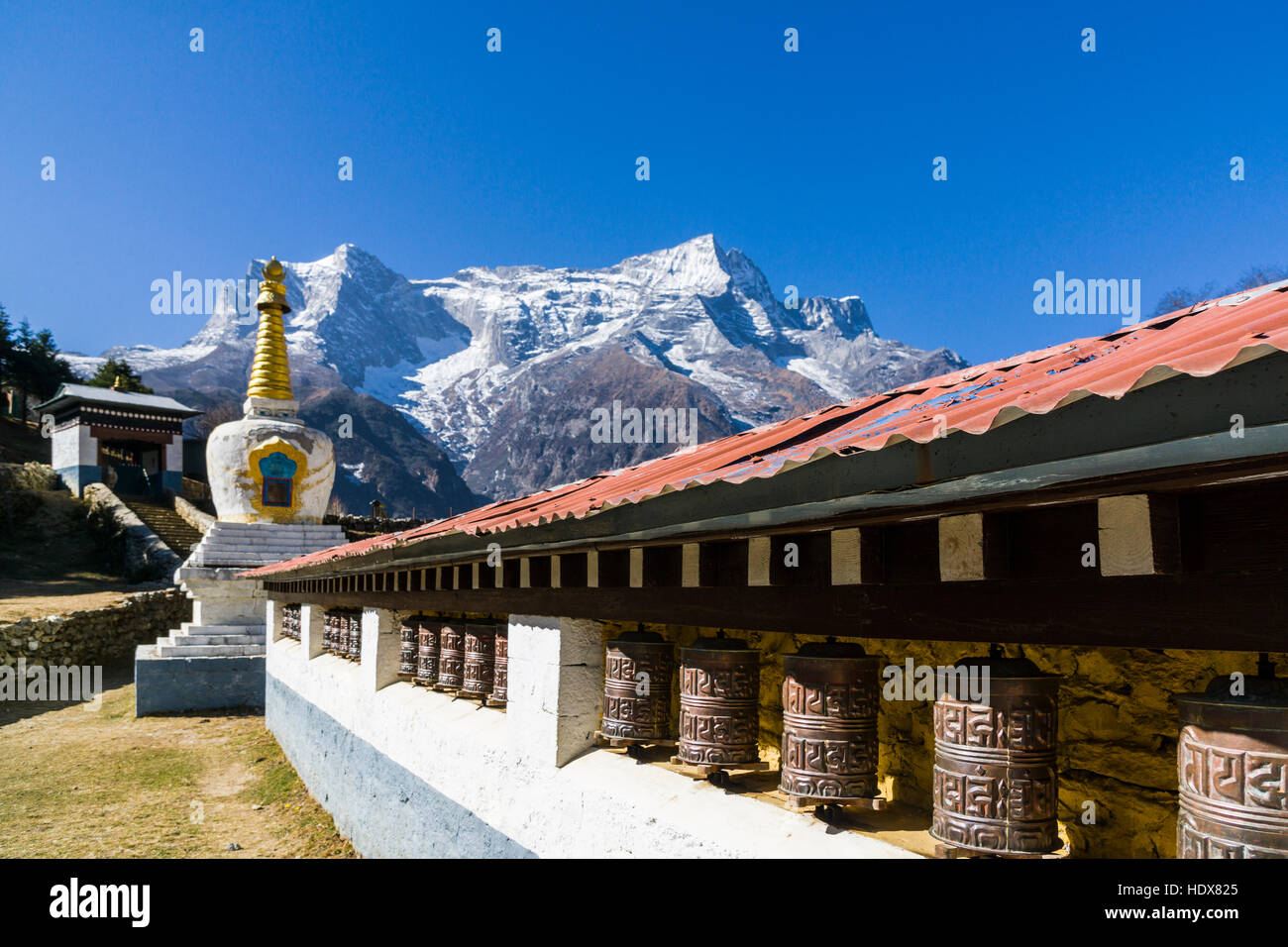 Una hilera de ruedas de oración tibetano y un blanco stupa, montañas cubiertas de nieve en la distancia Foto de stock