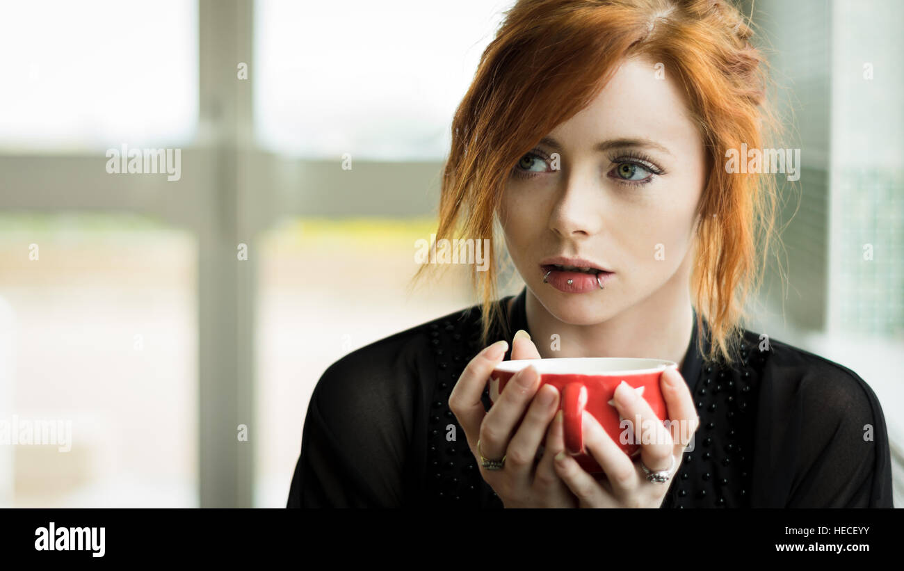 Pelirroja mujer con piercings bebiendo café desde una taza roja Foto de stock