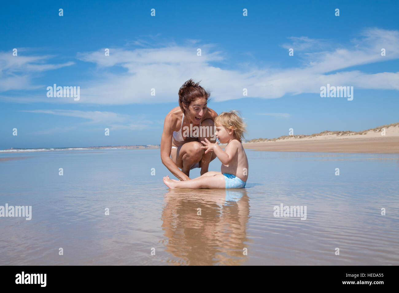 Familia de verano de dos años rubia con azul bañador bebé sentado sobre el  agua con una morena mujer madre en bikini blanco en cuclillas en la orilla  del mar playa Fotografía