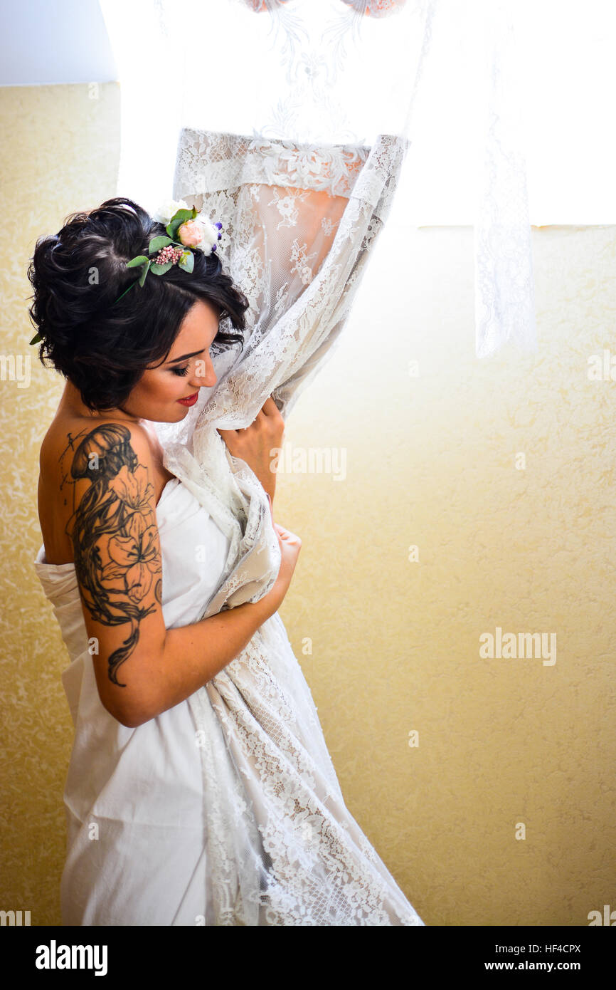 Hermosa morena sonriente posa con su novia boda vestido de encaje en la mano y mirando en el espejo. Vestida emocional flores blancas en el cabello. Foto de stock