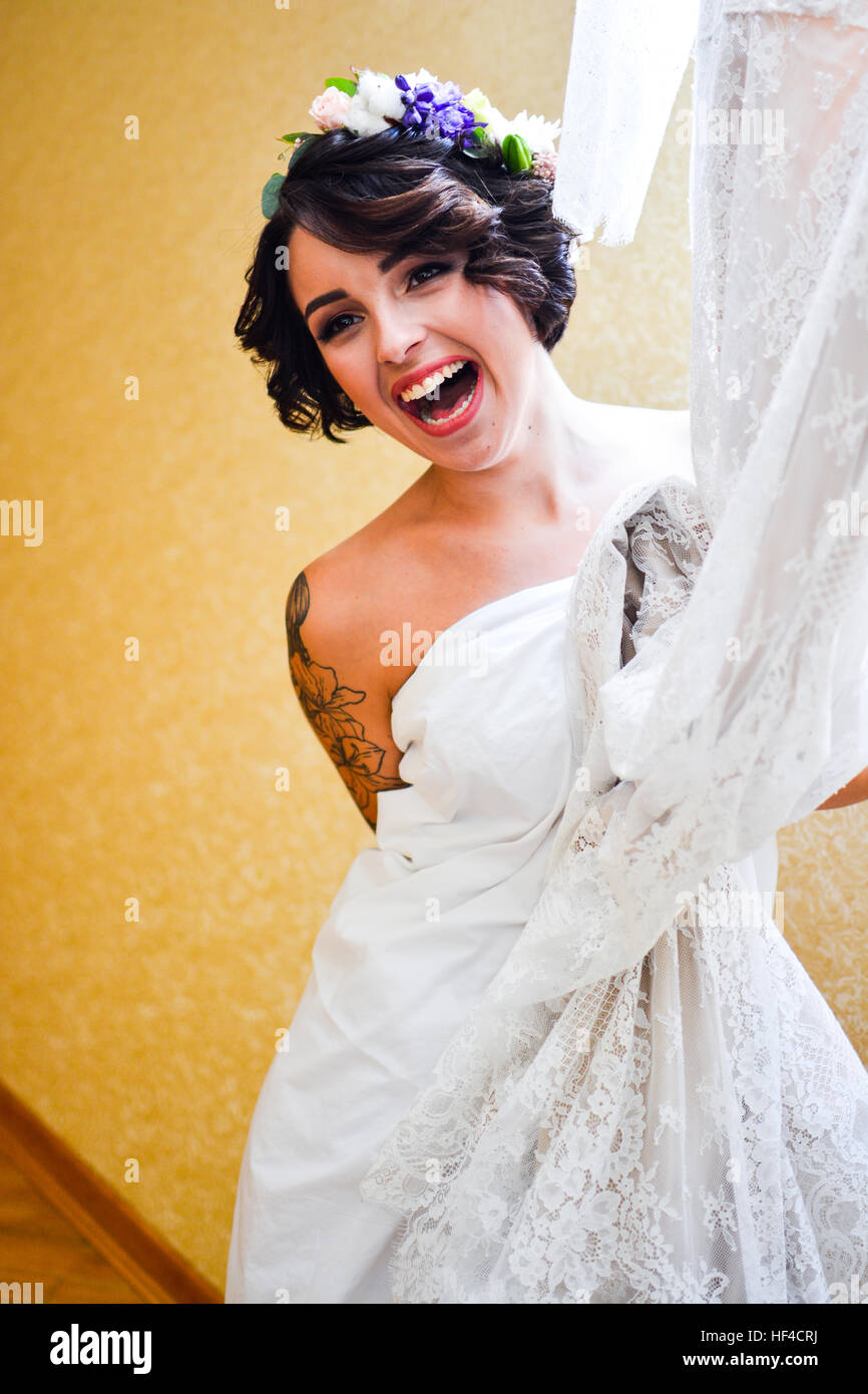 Hermosa morena sonriente posa con su novia boda vestido de encaje en la mano y mirando en el espejo. Vestida emocional flores blancas en el cabello. Foto de stock