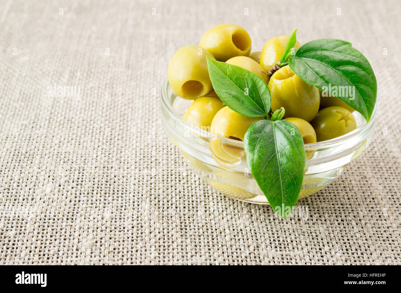 Aceitunas verdes con hojas verdes en un pequeño recipiente de cristal sobre un fondo de arpillera. Foto de stock