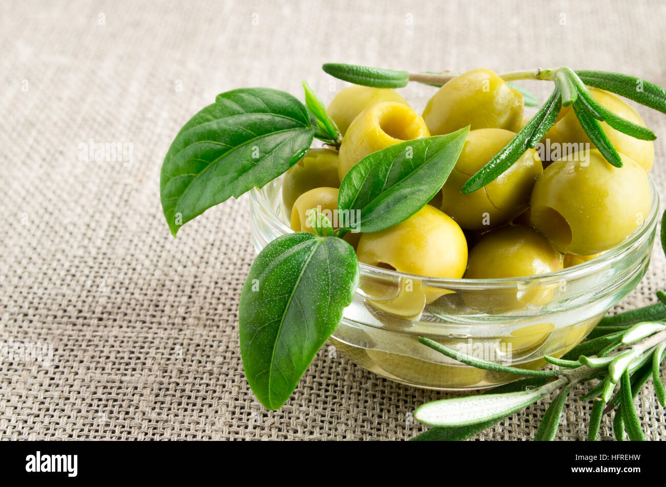 Aceitunas picadas con hojas verdes y el romero en un pequeño vaso en un viejo mantel Foto de stock