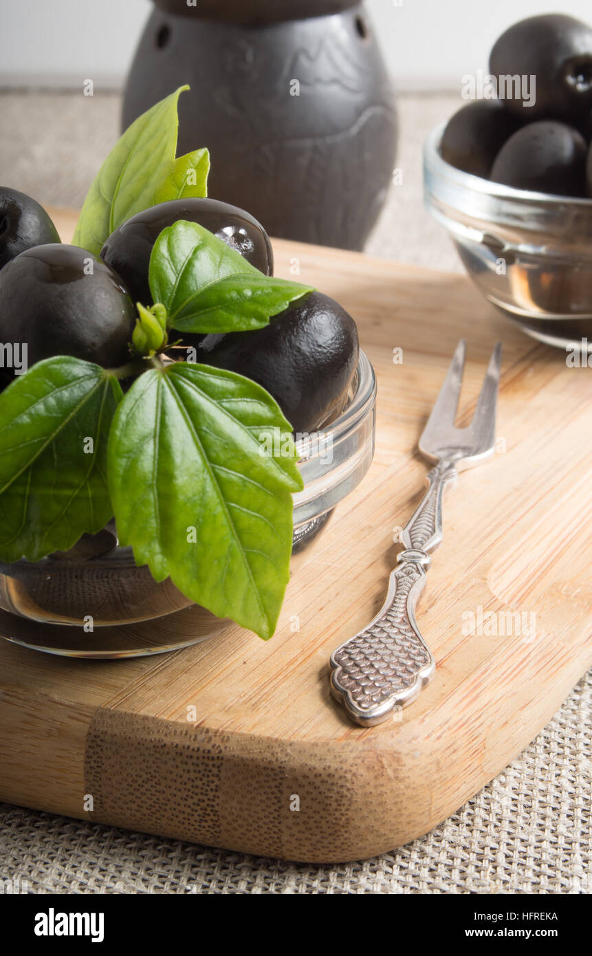 Aceitunas negras sobre una tabla de madera decorada con hojas verdes en el viejo vintage manteles y horquilla de metal Foto de stock