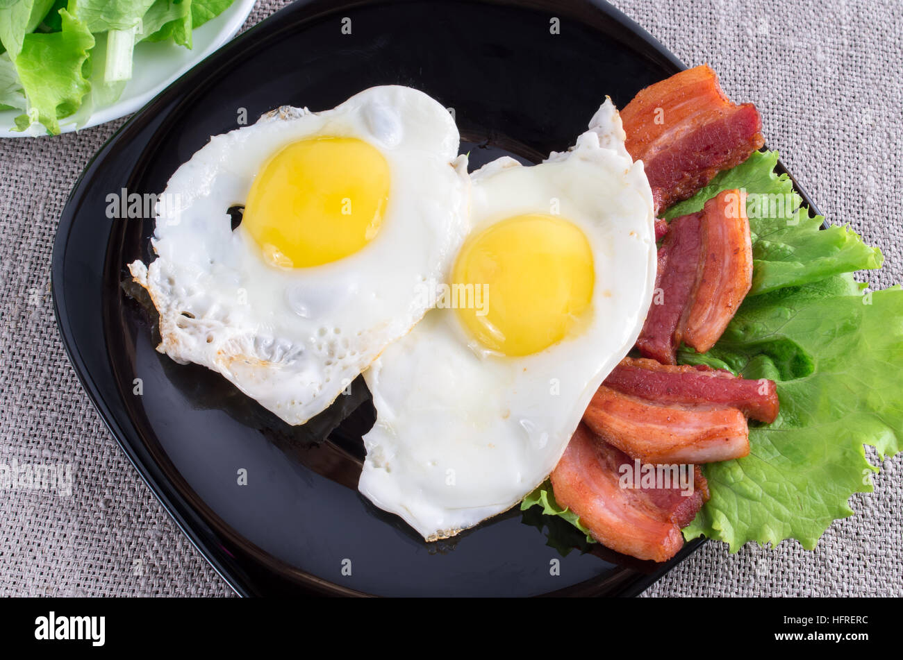 Vista superior de la placa negra con huevos fritos, tocino y lechuga de cerca en un mantel gris Foto de stock