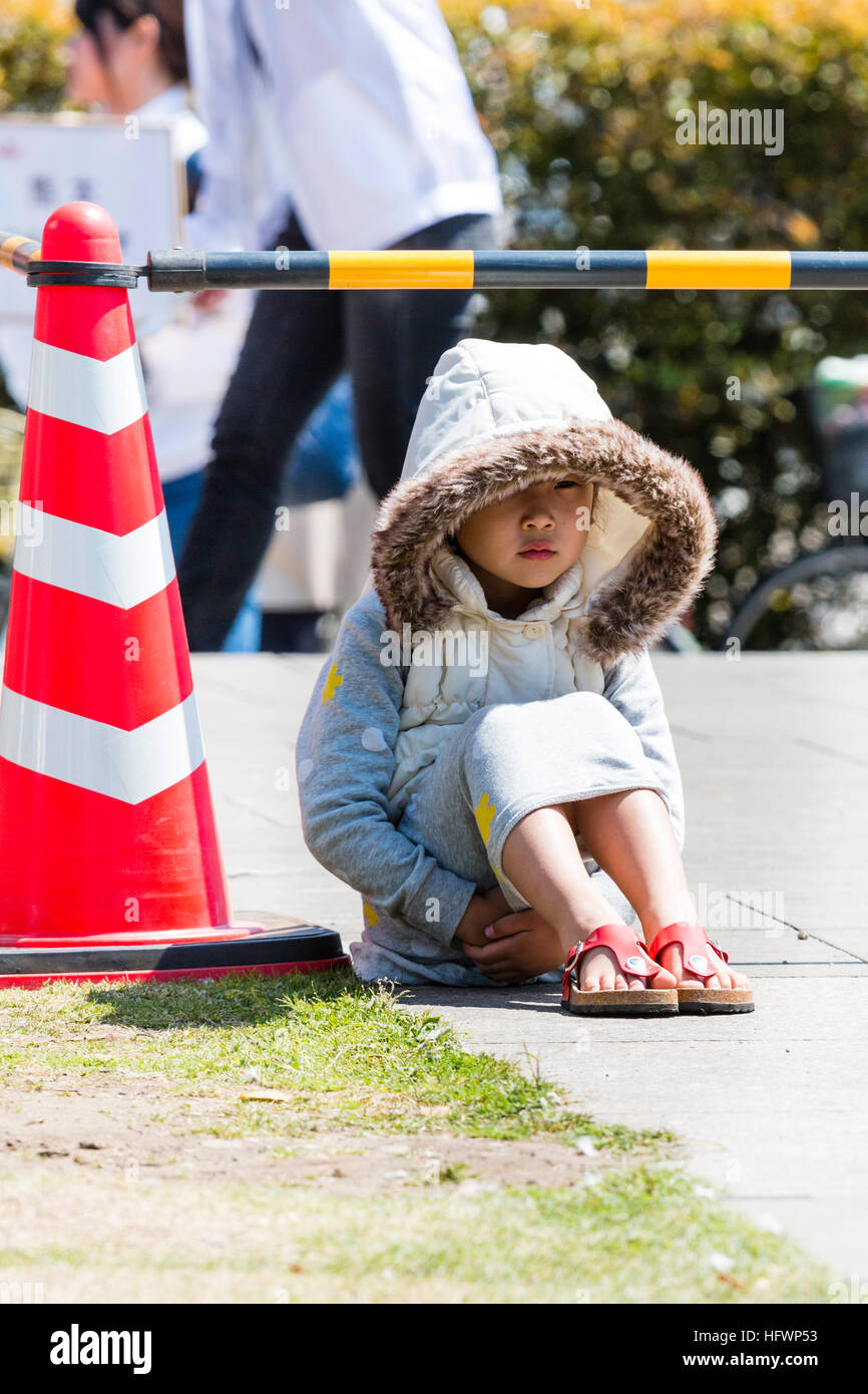 Japón, Kumamoto. Niño, Niña japonesa sentada en sol brillante de gran tamaño con capa y capucha, las manos entrelazadas bajo las piernas. Mirando Foto de stock