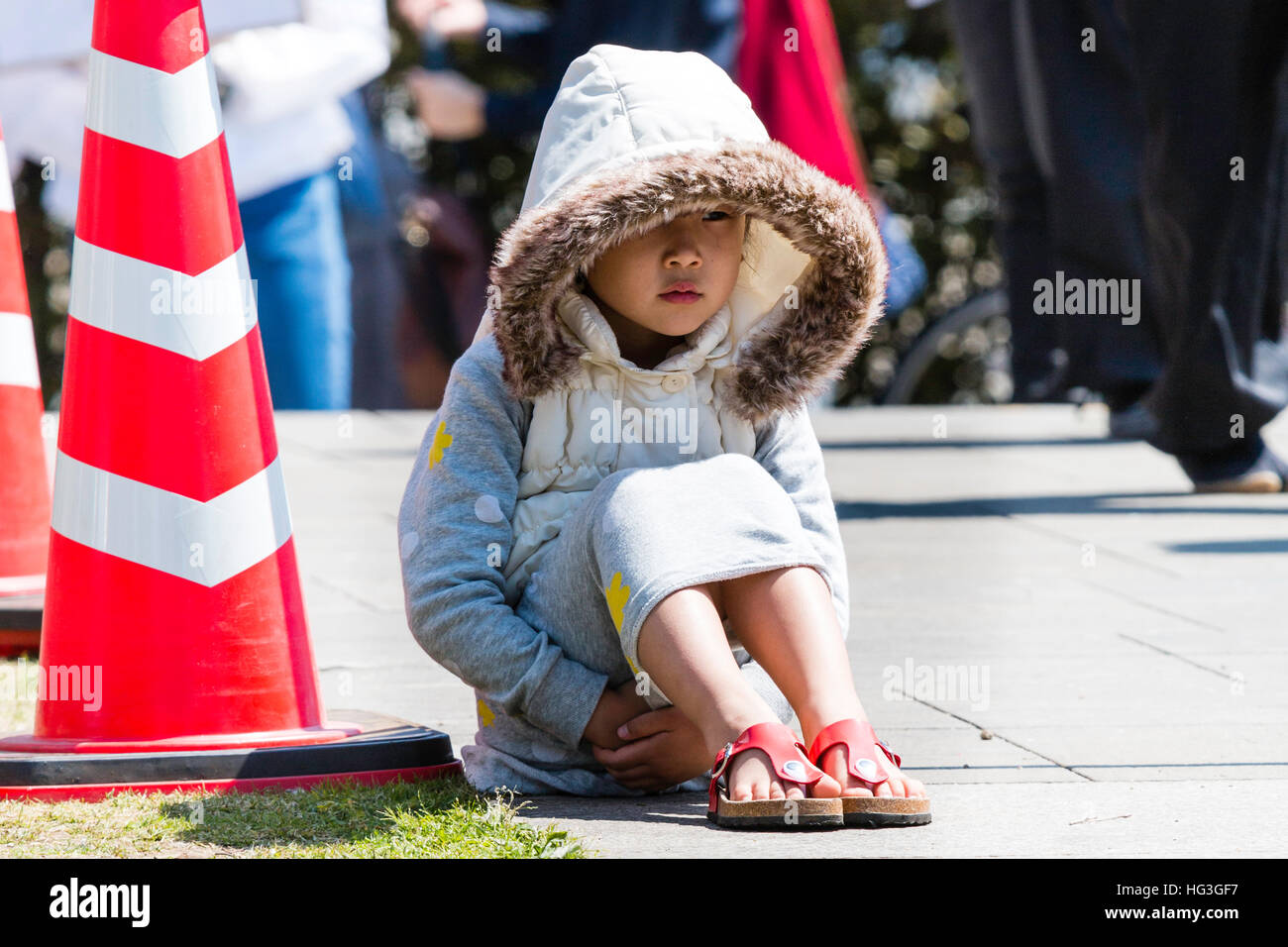 Japón, Kumamoto. Niño, Niña japonesa sentada en sol brillante de gran tamaño con capa y capucha, las manos entrelazadas bajo las piernas. Mirando Foto de stock