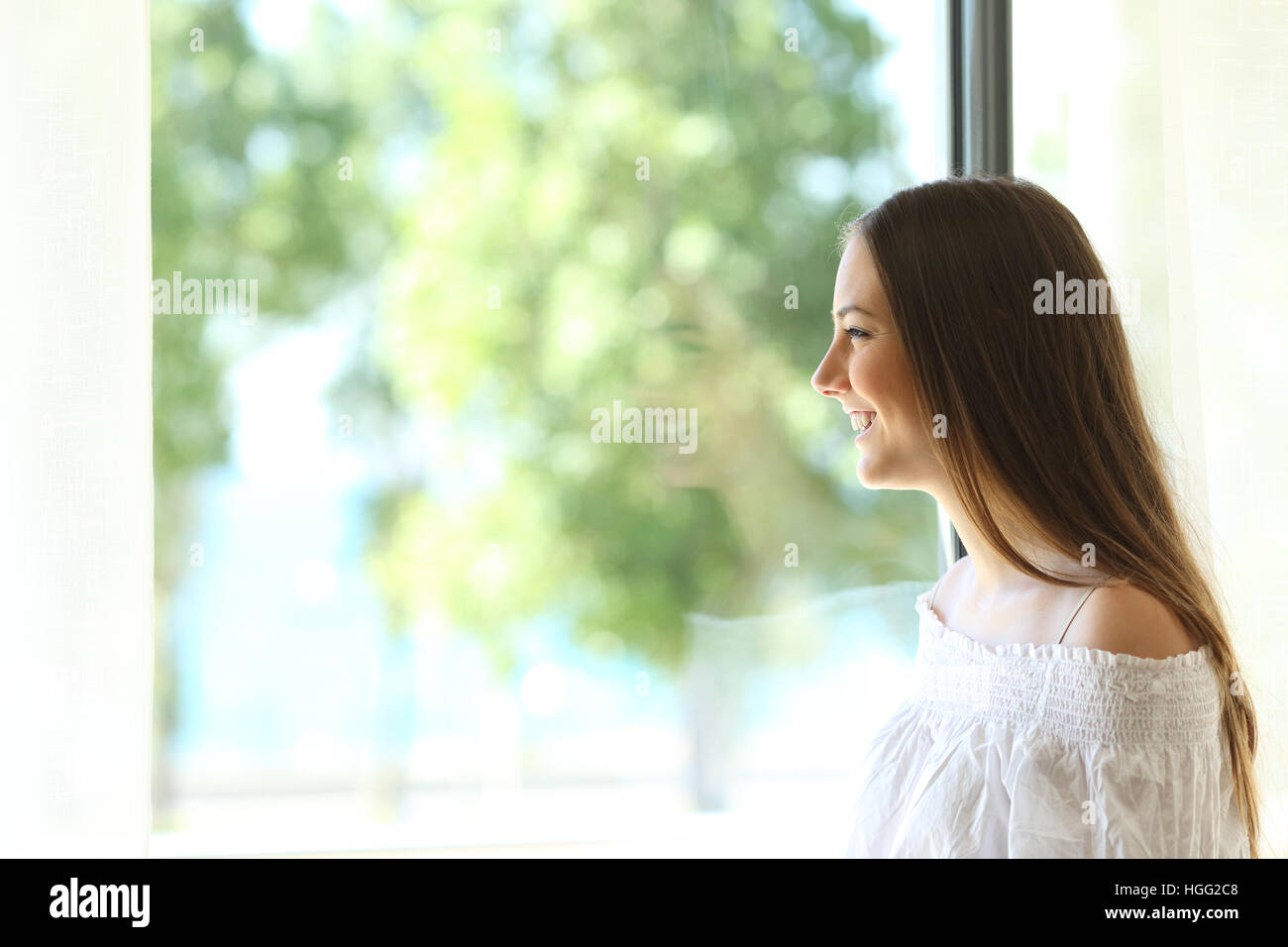 Vista lateral de una señora feliz en casa mirando afuera a través de una ventana de un salón o el dormitorio con un soleado fondo verde Foto de stock