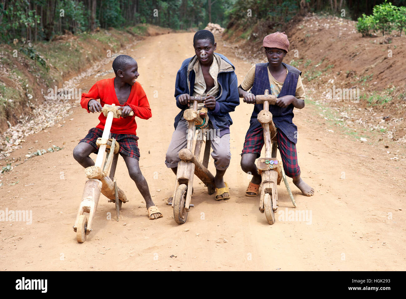 Tres niños ugandeses posan para una fotografía sobre sus bicicletas de madera en el África rural. Foto de stock