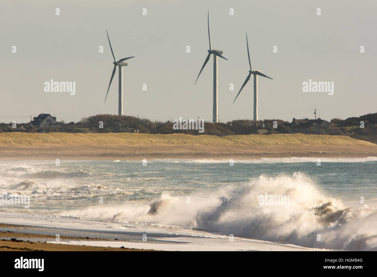 Los aerogeneradores de un parque eólico stand junto a la arena de la costa de Wexford en Irlanda con potentes olas que rompen en la orilla Foto de stock