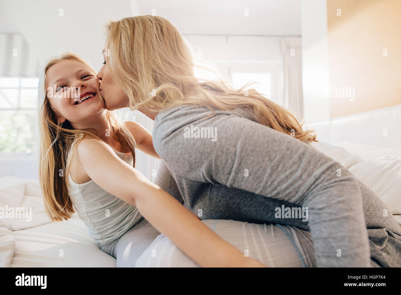 Mujer joven besos niña mientras está sentado en el dormitorio. Amorosa madre e hija en la cama. Foto de stock