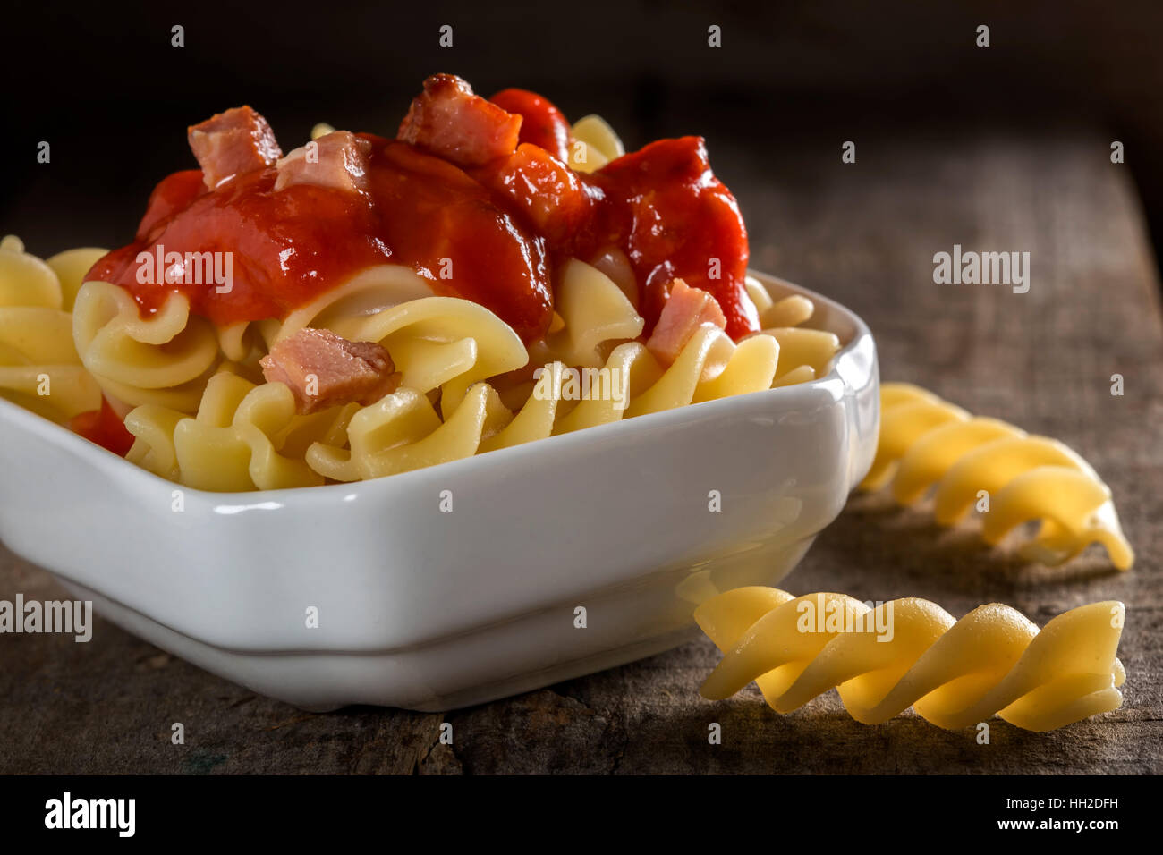 Fusilli pasta con salsa de tomate y bacon frito en tazón blanco sobre fondo de madera Foto de stock