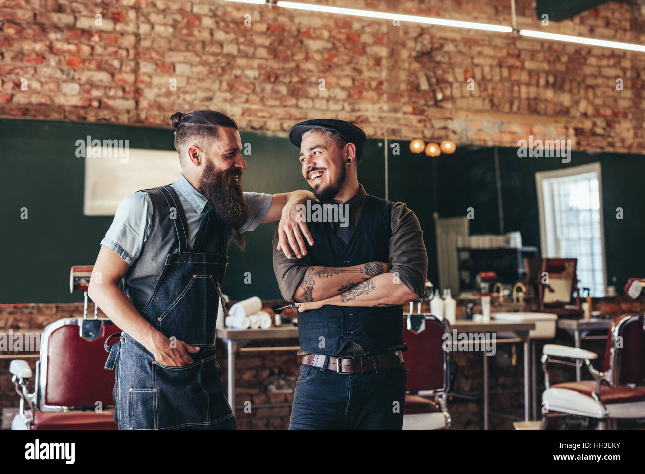 Foto de feliz barbero con el cliente de pie en el barbershop y sonriente. Dos hombre en el salón, el peluquero y el cliente. Foto de stock