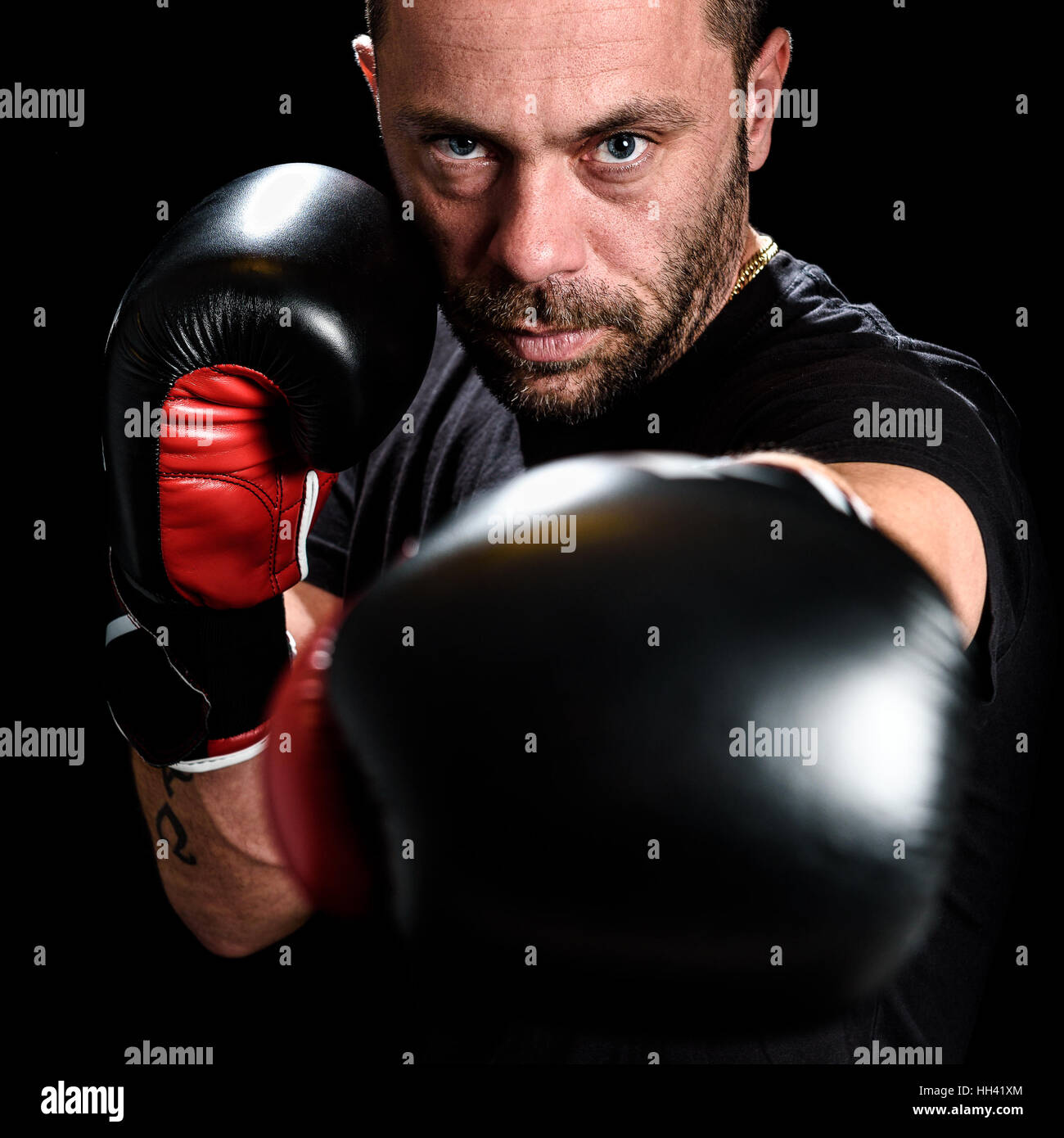 Retrato de atleta masculino boxer hombre mirando agresivo con guantes de boxeo, camisa negra y tatuajes. Aislado sobre fondo negro Foto de stock