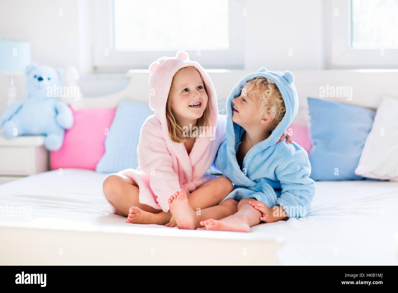 Feliz riendo los niños, niño y niña en suave albornoz después baño juego de cama de color blanco con azul y rosa almohadas en la soleada habitación Fotografía de stock -