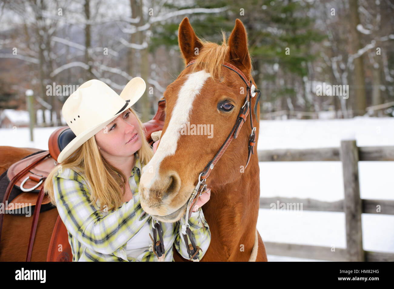 Cowgirl blanco con sombrero de cowboy, vistiendo trajes de equitación occidental mirando un caballo con afecto en un cercado de pastos de invierno Foto de stock