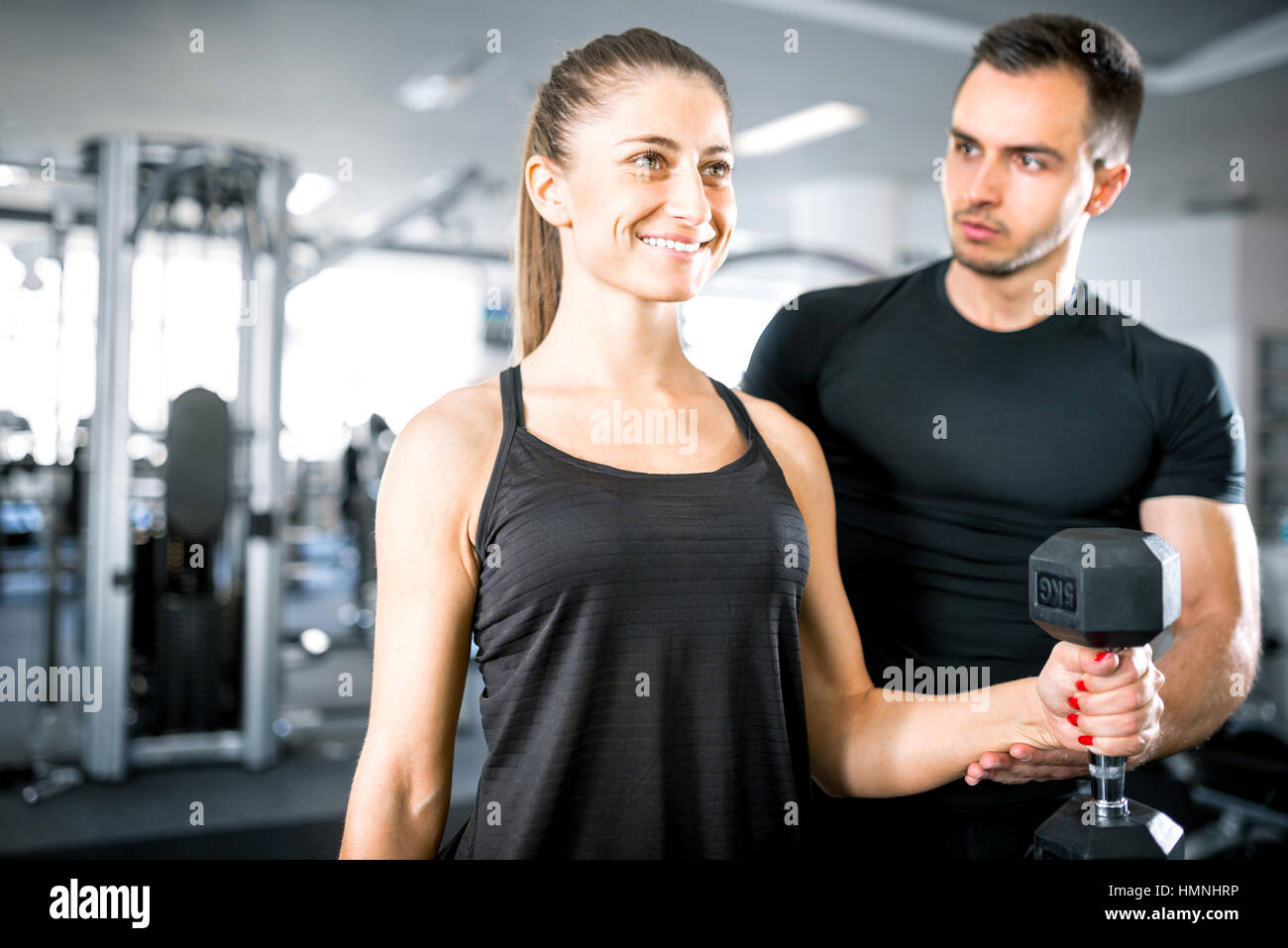 Adulto joven mujer trabajando en el gimnasio, haciendo flexiones de brazos (bíceps) con la ayuda de su entrenador personal. Foto de stock