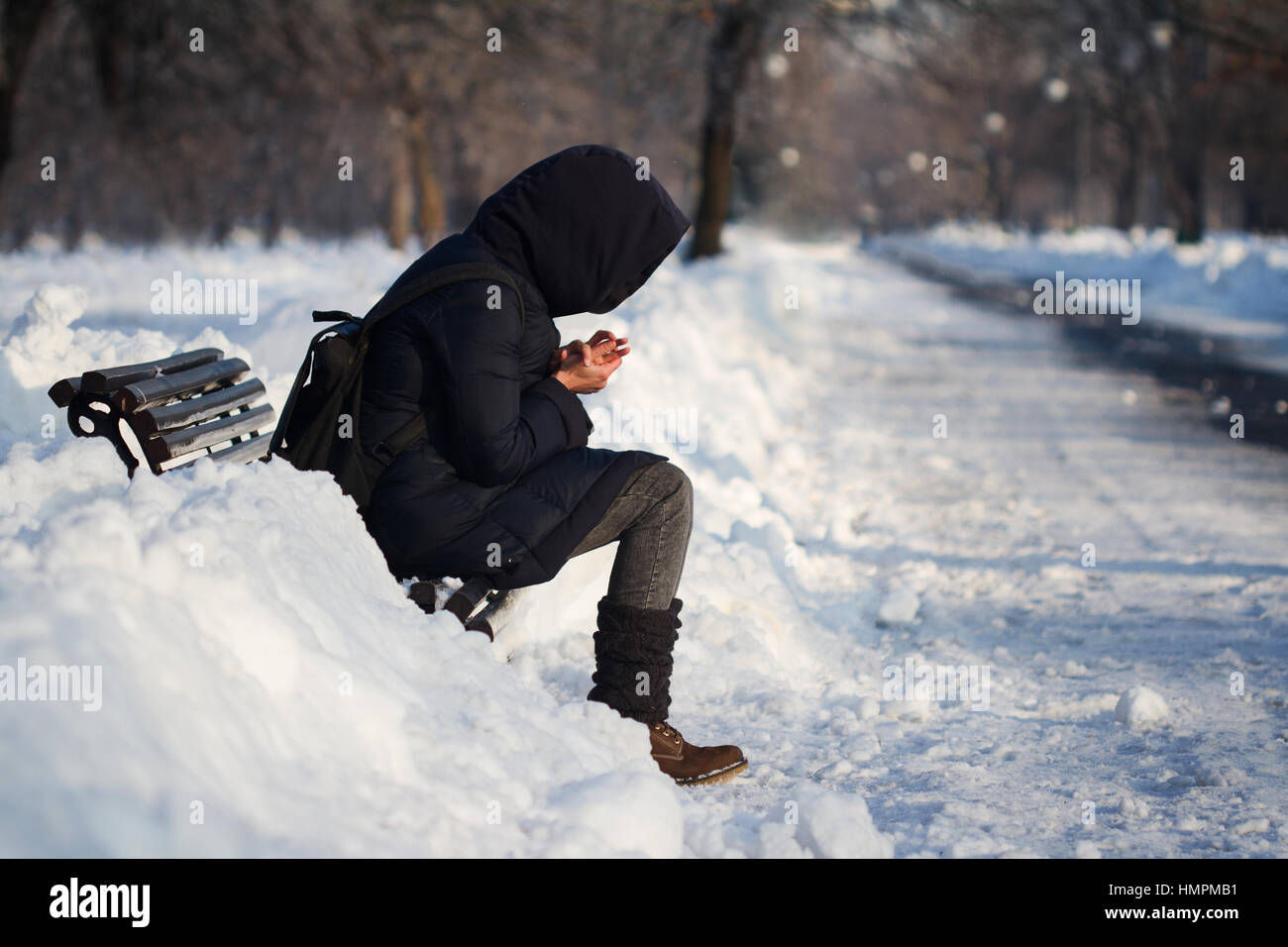 Persona solitaria con mochila hoodie y alojarse en un banco en el parque de la ciudad callejón con nieve en invierno y el frío Foto de stock