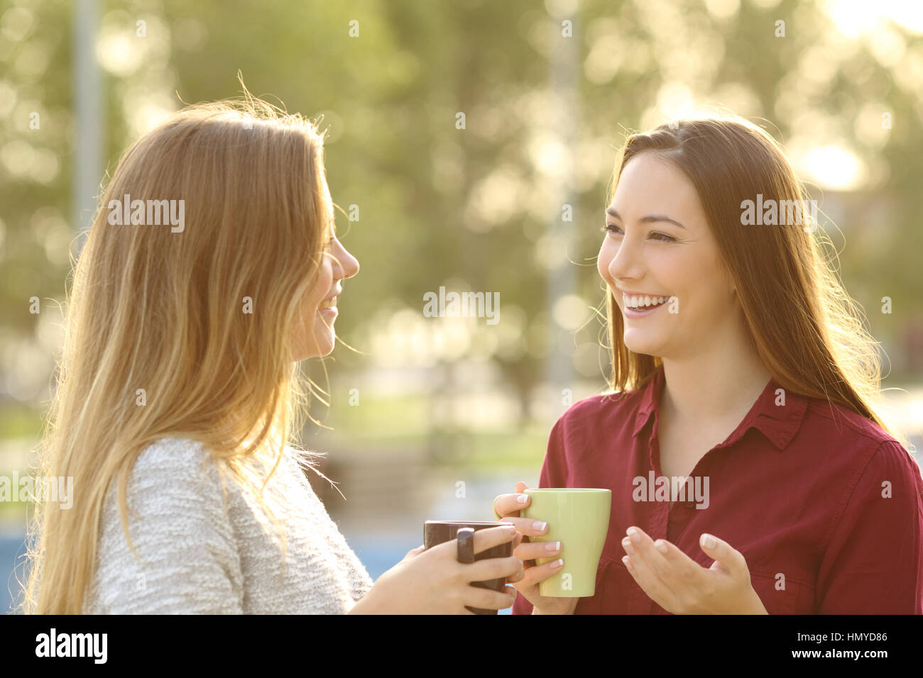 Dos amigos hablando feliz celebración taza de café al aire libre en un parque con un fondo verde al atardecer con una cálida luz trasera Foto de stock