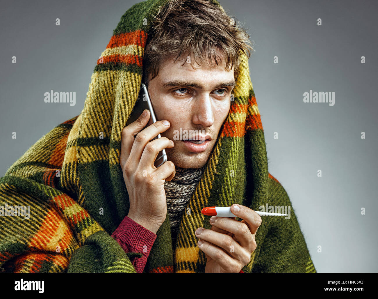 Hombre enfermo envuelto en una manta con una temperatura alta llamando al teléfono. Hombre que sufría del virus de la gripe y frío invierno. Concepto de salud Foto de stock