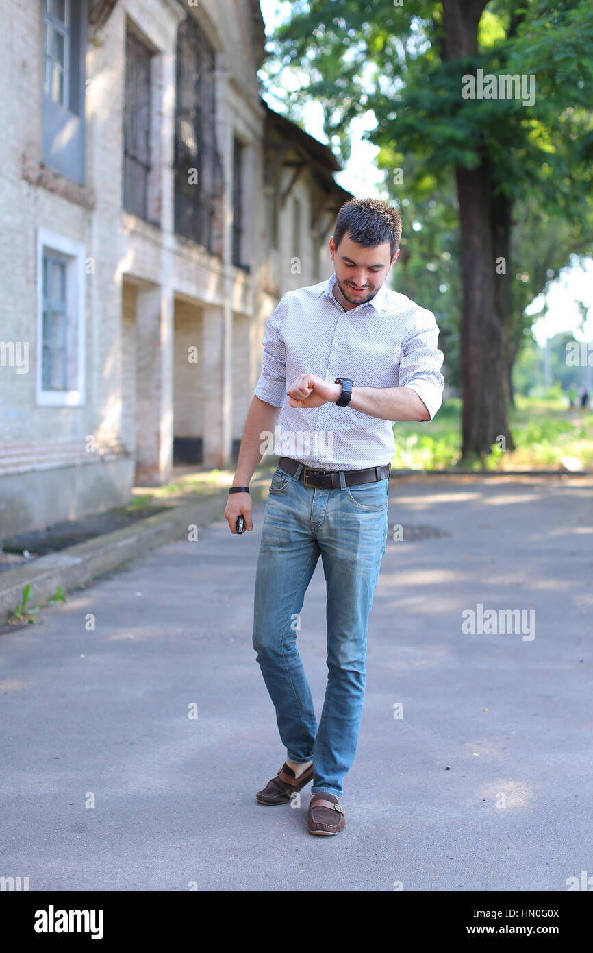 Joven barbudo hombre hombre hombre va caminando a lo largo de la carretera  y mirando a ver en el brazo. Joven vestida con la camisa blanca con rayas  rojas, blue jeans y