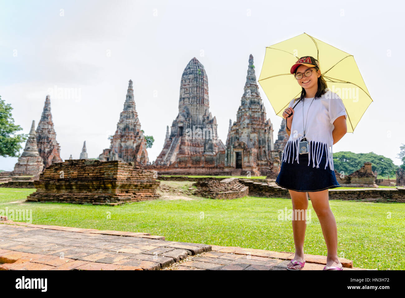 Turista lindo adolescente con gafas con una cámara en el cuello, mirando y sonrió felizmente sosteniendo el paraguas en el templo Wat Chaiwatthanaram antecedentes Foto de stock