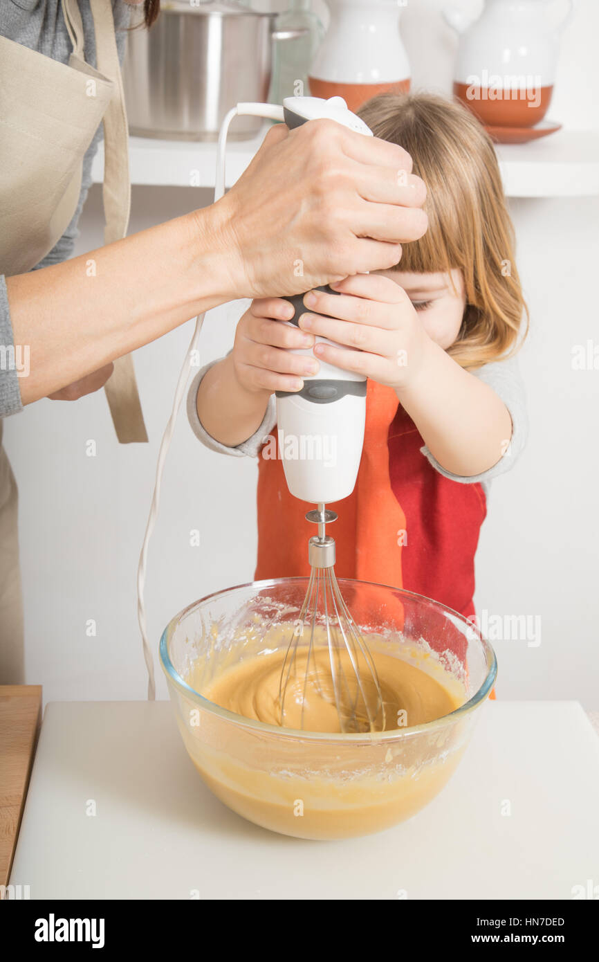 Tres años de edad, la crema en el tazón de vidrio con batidora eléctrica, y  la mujer madre en el trabajo en equipo para hacer un bizcocho y cocinar en  la cocina
