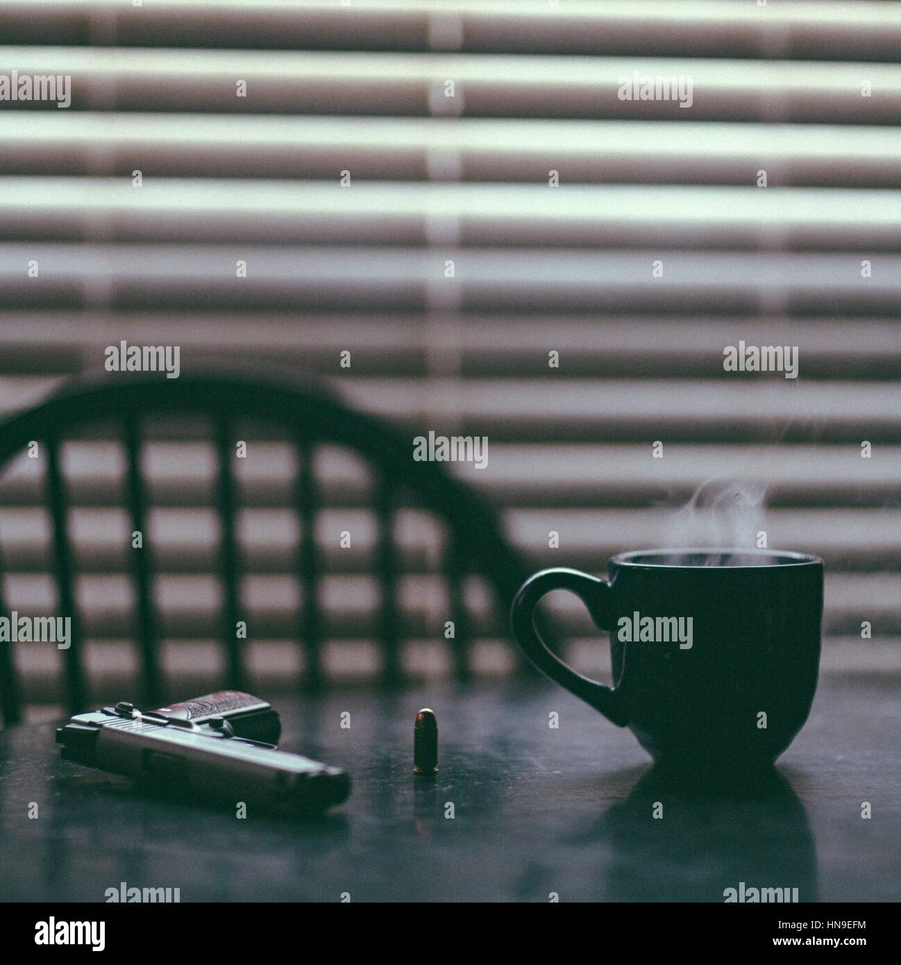Una escena ominosa/vida fija de una mesa que sostiene una taza caliente de café, una pistola, y una sola bala. En el fondo hay una sola silla y persianas cerradas Foto de stock