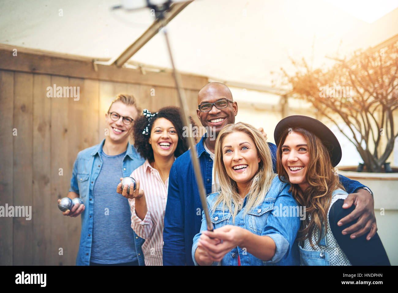 Cinco amigos jóvenes, hombres y mujeres, teniendo selfie con selfie stick celebración petanca bolas de metal en las manos, sonriendo y mirando hacia arriba Foto de stock