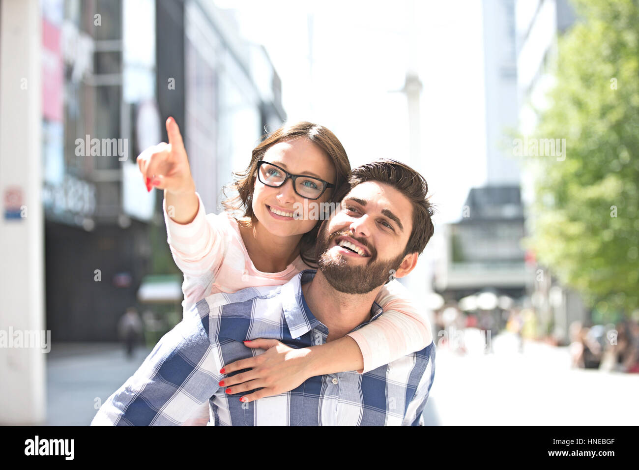 Mujer alegre apuntando hacia fuera mientras disfruta piggyback paseo sobre el hombre en la ciudad Foto de stock