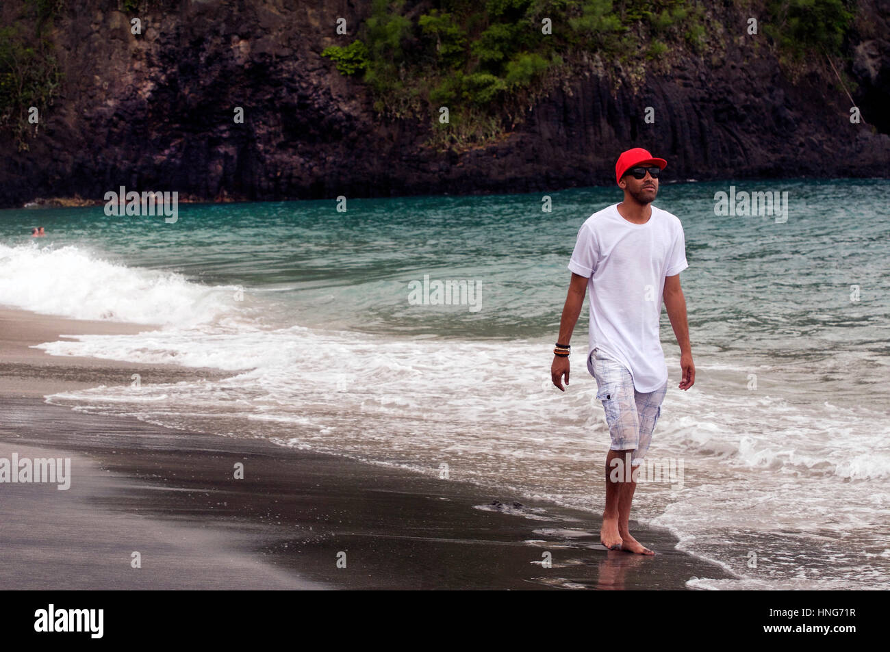 Cool Dude paseos a lo largo de Playa en Bali Indonesia vestidos de rojo Baseball Hat, camiseta blanca y pantalón corto Foto de stock