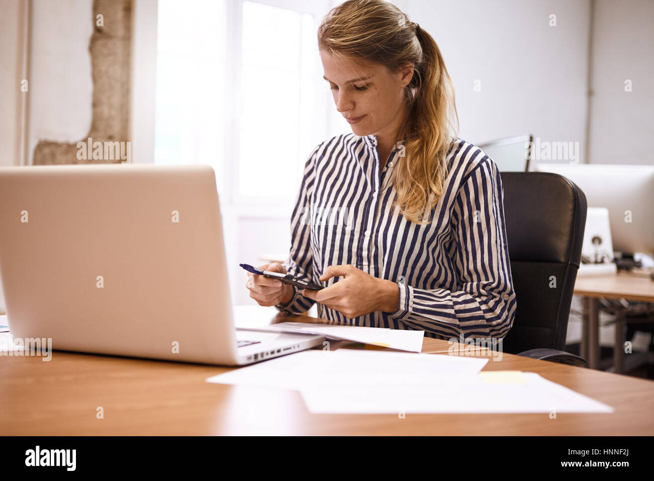 Empresa joven mujer haciendo cálculos en su teléfono mientras está sentado  en su escritorio con un ordenador portátil y varias notas en frente de ella  Fotografía de stock - Alamy
