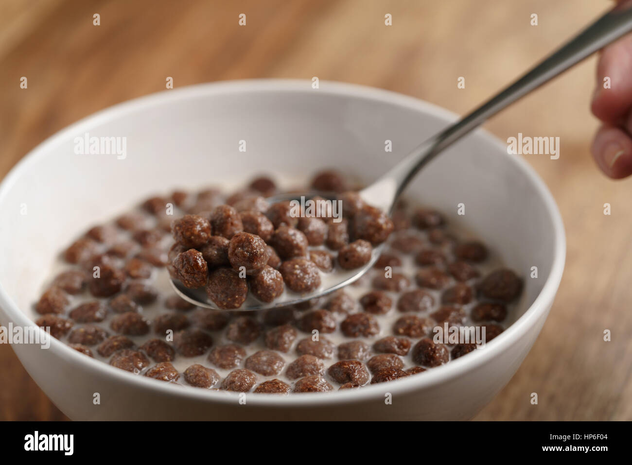 Comer con cuchara bolas de cereales de chocolate con leche en el recipiente blanco para desayunar en la mesa de madera, enfoque superficial Foto de stock