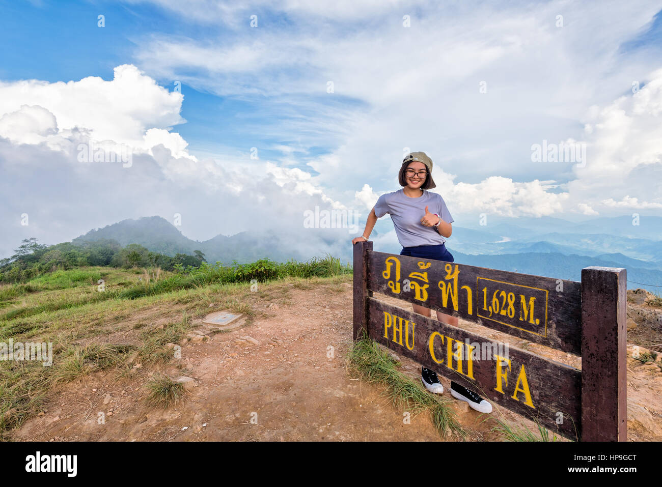 Turista jovencita excursionista tapa de desgaste y gafas sonrisa permanente plantean pulgar arriba, cerca de las atracciones de placa de Phu Chi Fa Forest Park en la montaña Foto de stock