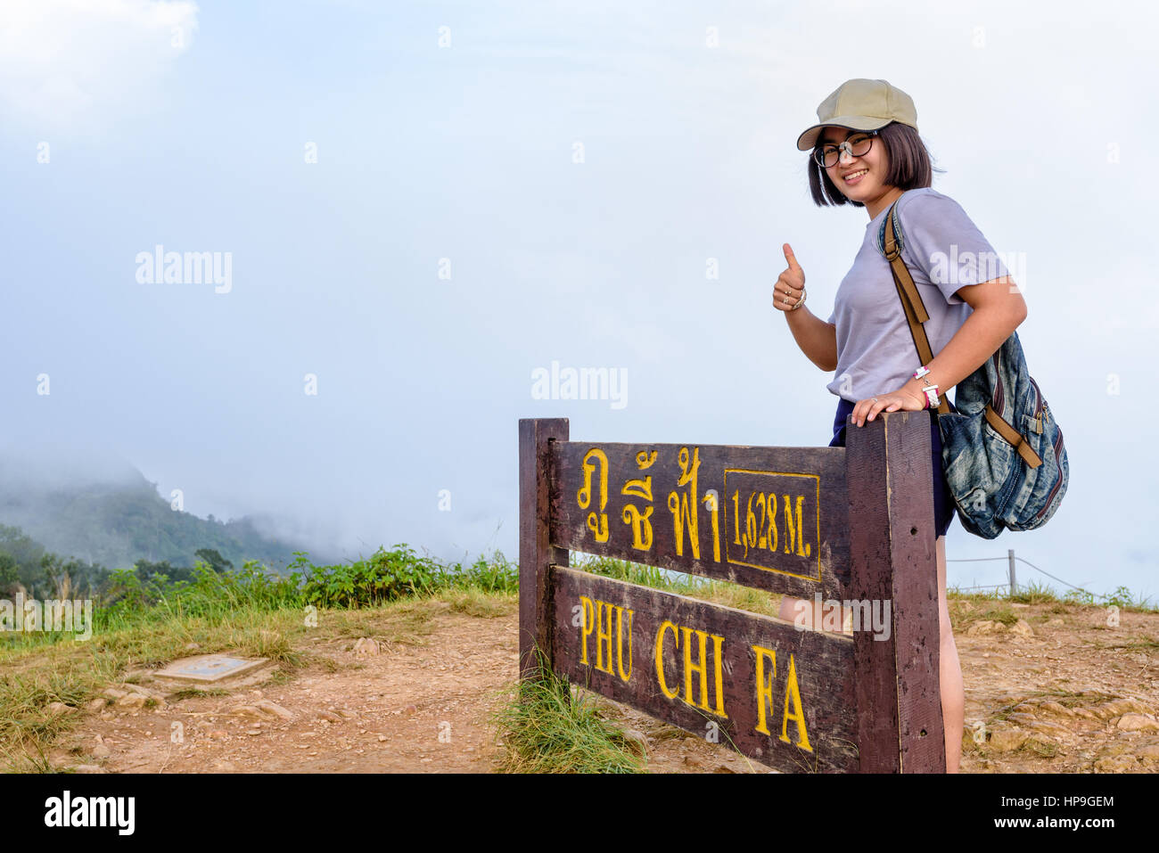 Turista jovencita excursionista tapa de desgaste y gafas con mochila sonriendo pulgar arriba, cerca de las atracciones de placa de Phu Chi Fa Forest Park en montaña y niebla Foto de stock
