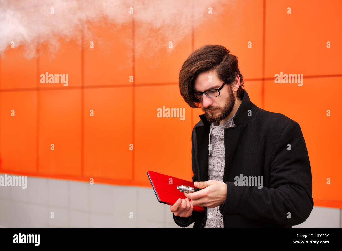 Young hipster hombre con barba y gafas fuma un cigarrillo electrónico. Él  sostiene un tablet pc. La moda juvenil. Delicious pares. Sin nicotina liqu  Fotografía de stock - Alamy