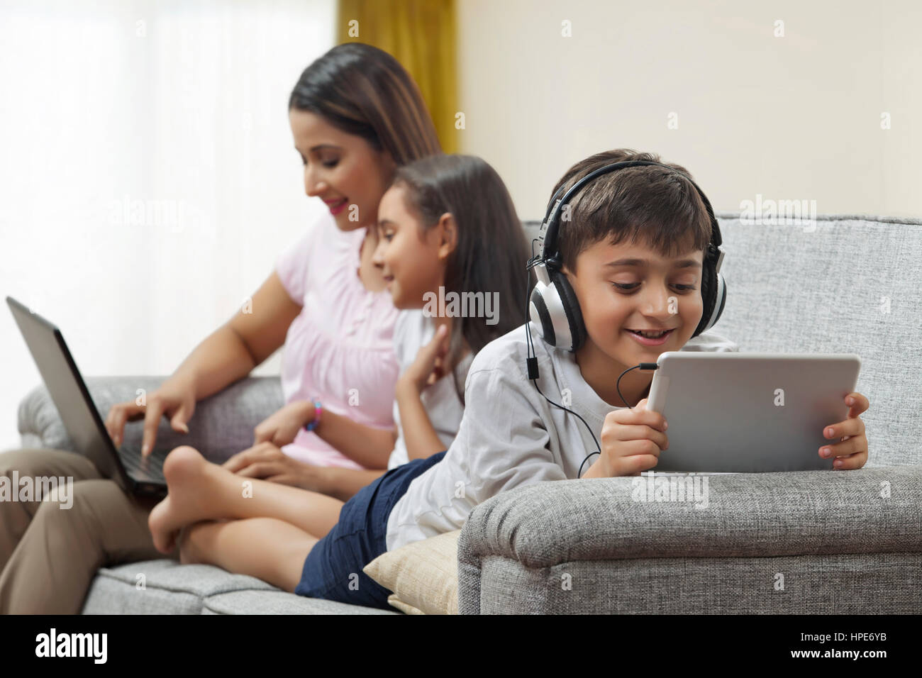 Centrado boy utilizando tablet digital auriculares con madre y hermana utilización portátil en segundo plano. Foto de stock