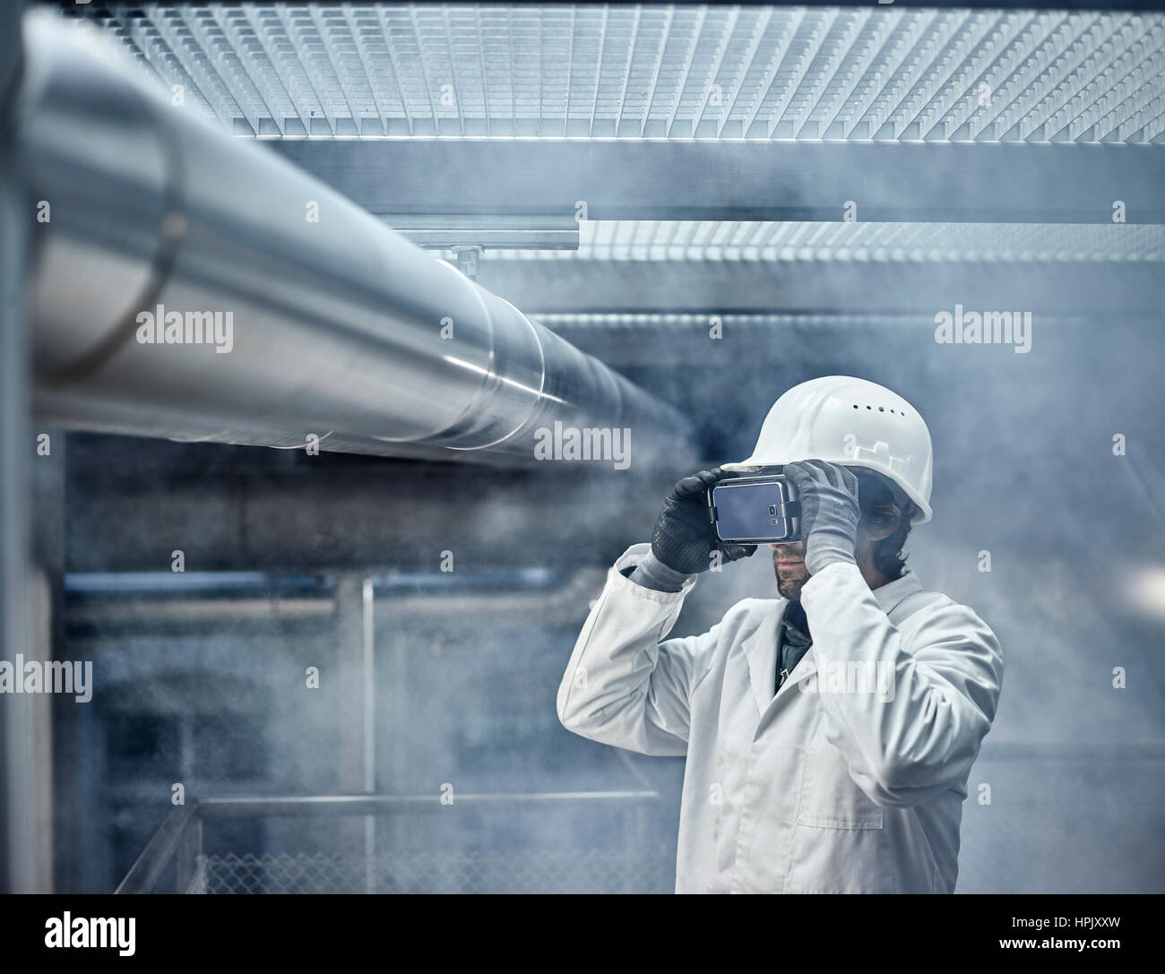 Hombre con gafas VR, casco blanco y bata de laboratorio de pie en frente de la planta industrial, Austria Foto de stock