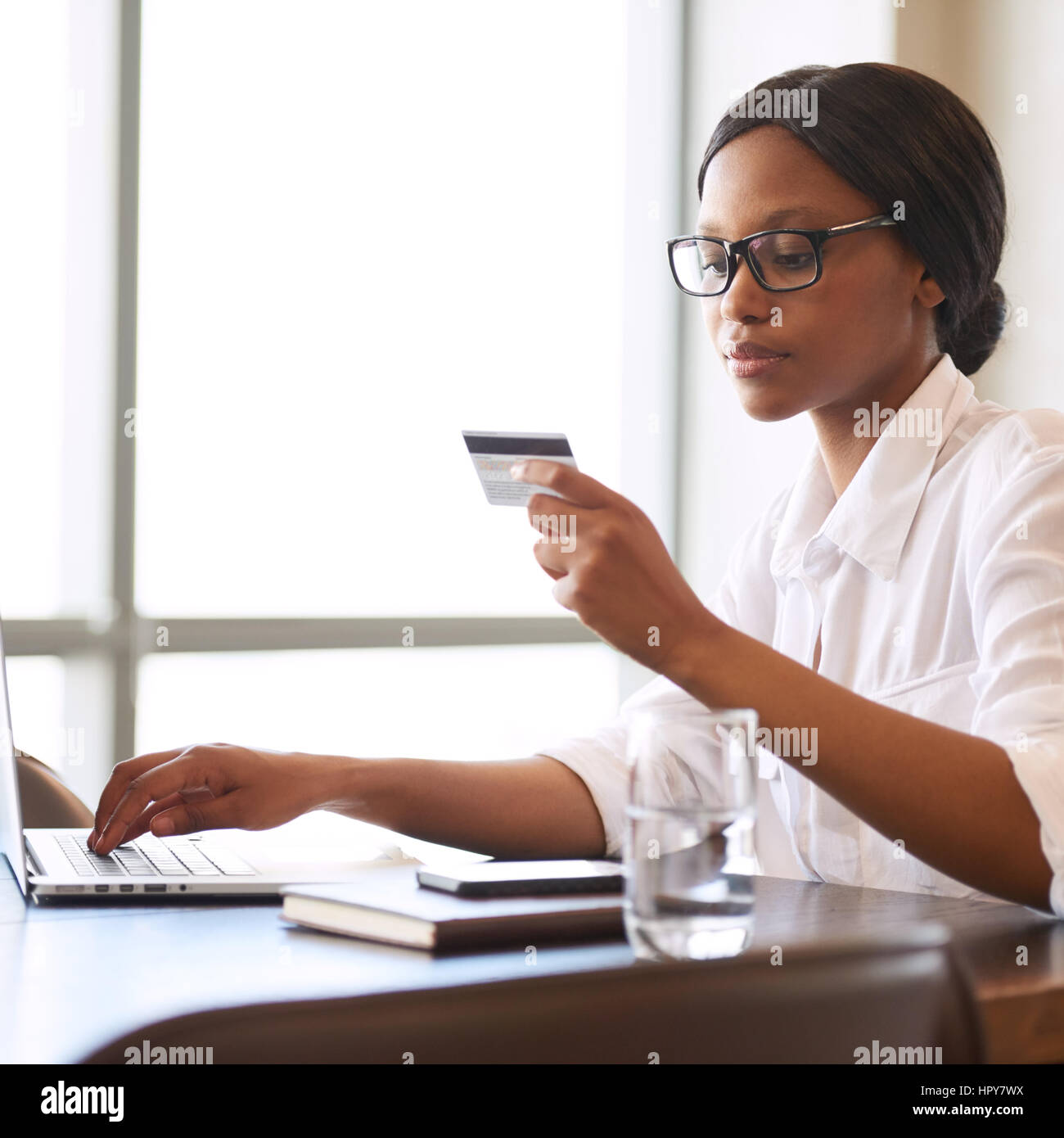 Imagen cuadrada de un bonito joven mujer negra ocupada la celebración de una tarjeta de crédito en una mano leyendo la información requerida y escribiendo en el pertinente di Foto de stock