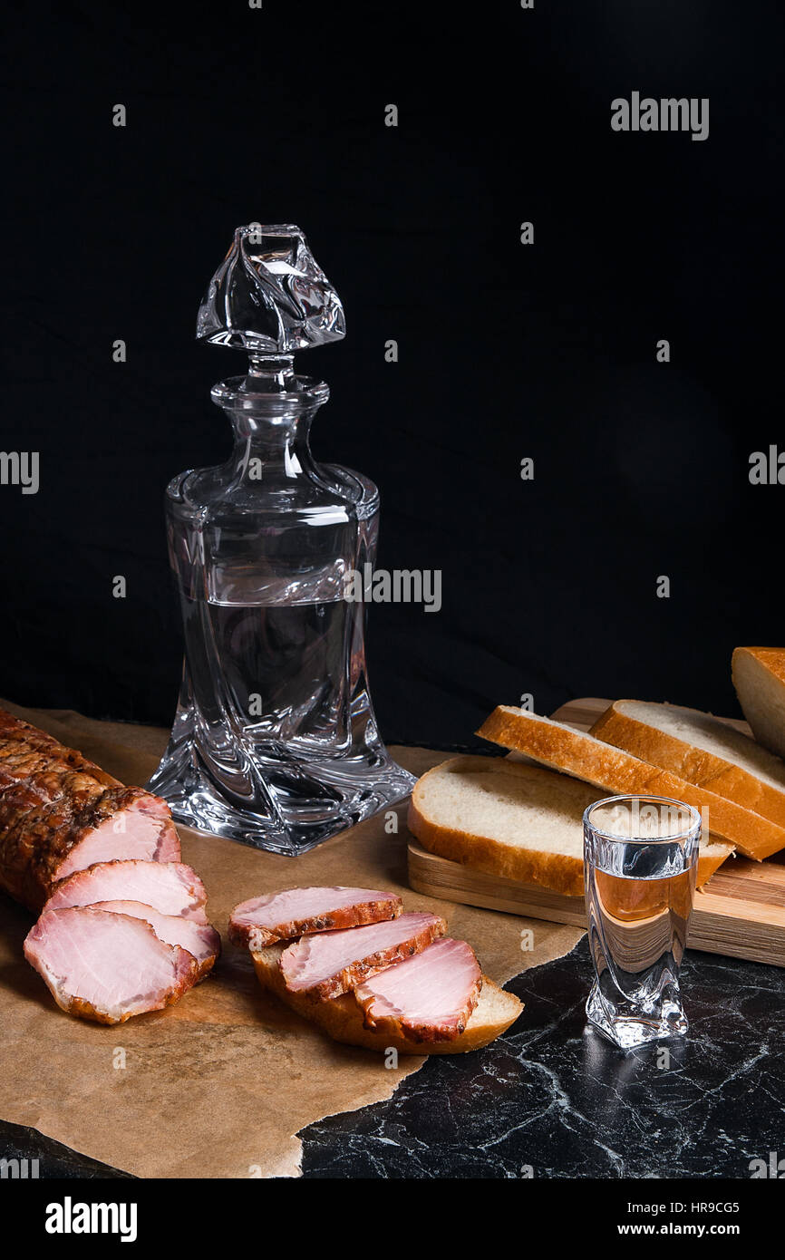 Botella y shot gafas con vodka. Un pequeño aperitivo de pan y carne cerca  del cristal del tiro. Cortes de carne ahumada o jamón en papel de embalar  marrón. Whea blanco Fotografía