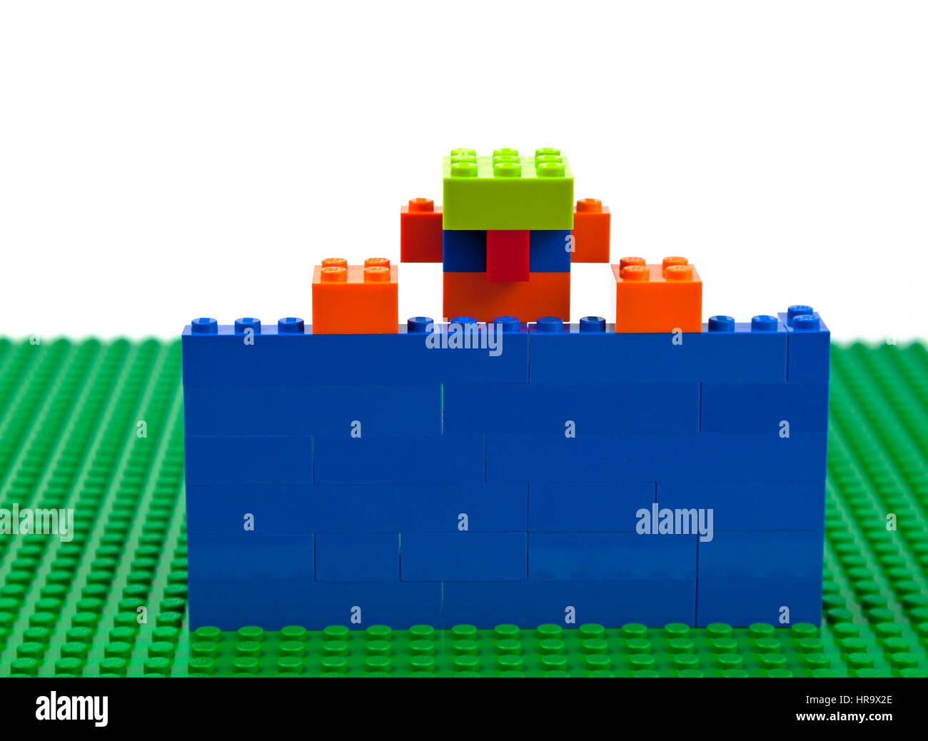 Construcción de ladrillos de Lego de un muchacho, hombre, macho con jarra-orejas y una gorra verde confinados detrás de una pared azul elevando a sí mismo a huir o simplemente decir "hola". Foto de stock