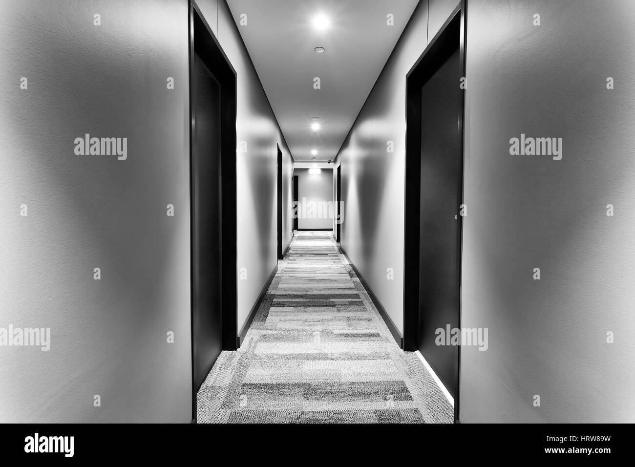 Corredor simétrico en el moderno y flamante hotel con habitaciones en los lados de las puertas y paredes blancas iluminadas por luces brillantes. No invitados a la vista. Foto de stock