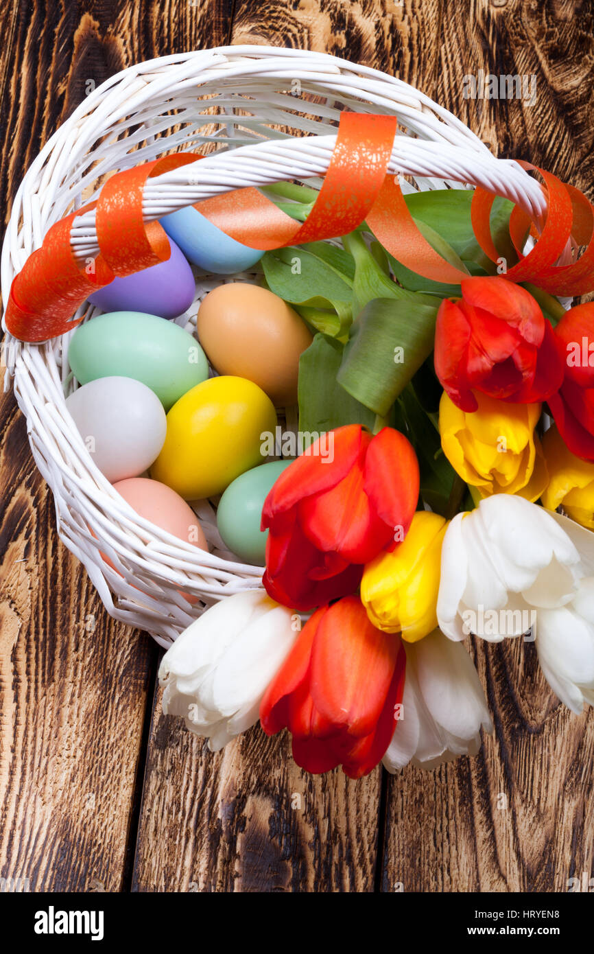 Los huevos de pascua en una cesta con coloridos tulipanes blancos sobre fondo de madera Foto de stock