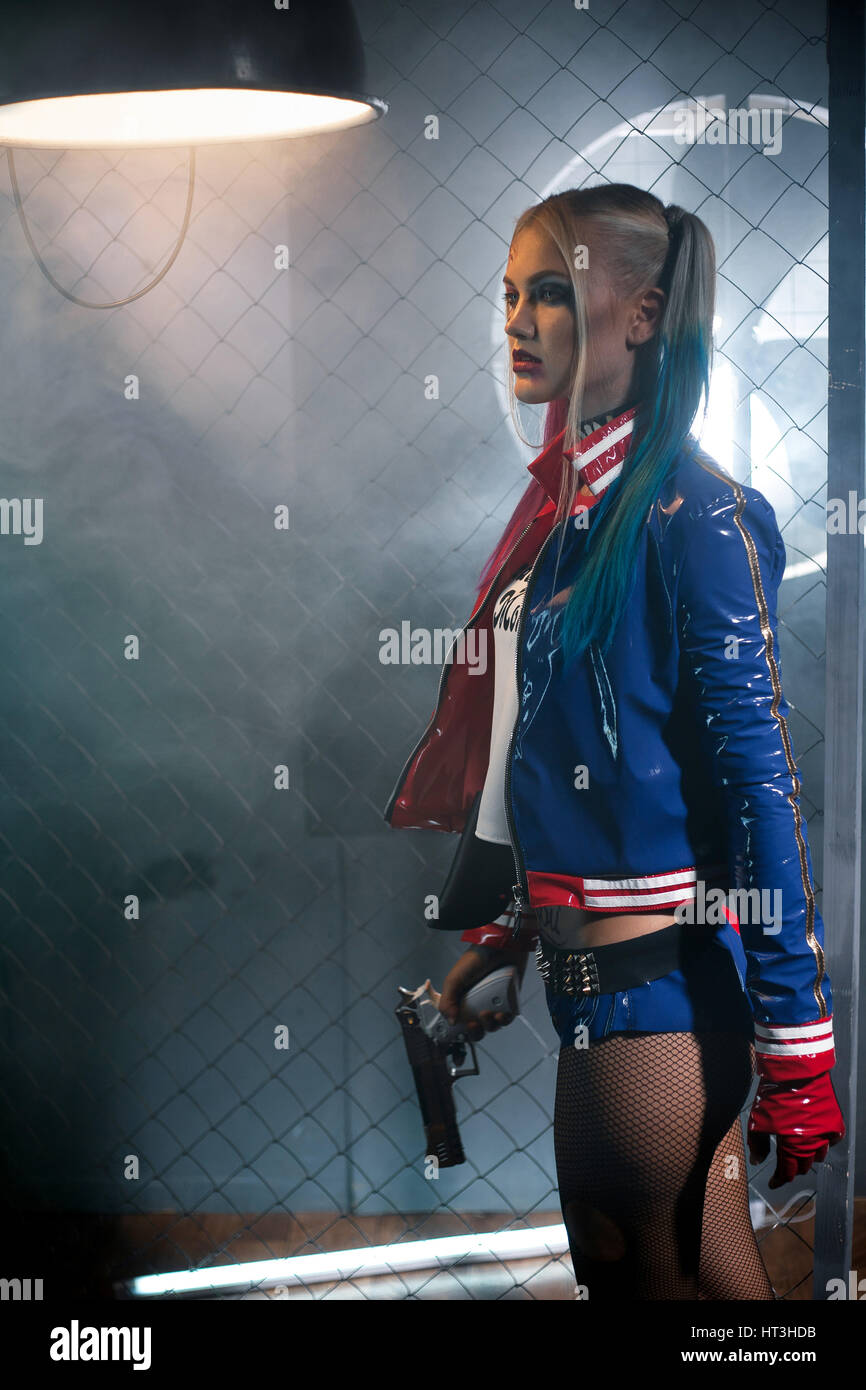 Pigmalión metálico recluta Chica en traje Harley Quinn con pistola. Cosplay. Ella representa cerca de  rejilla bajo la lámpara. Cosplay Fotografía de stock - Alamy