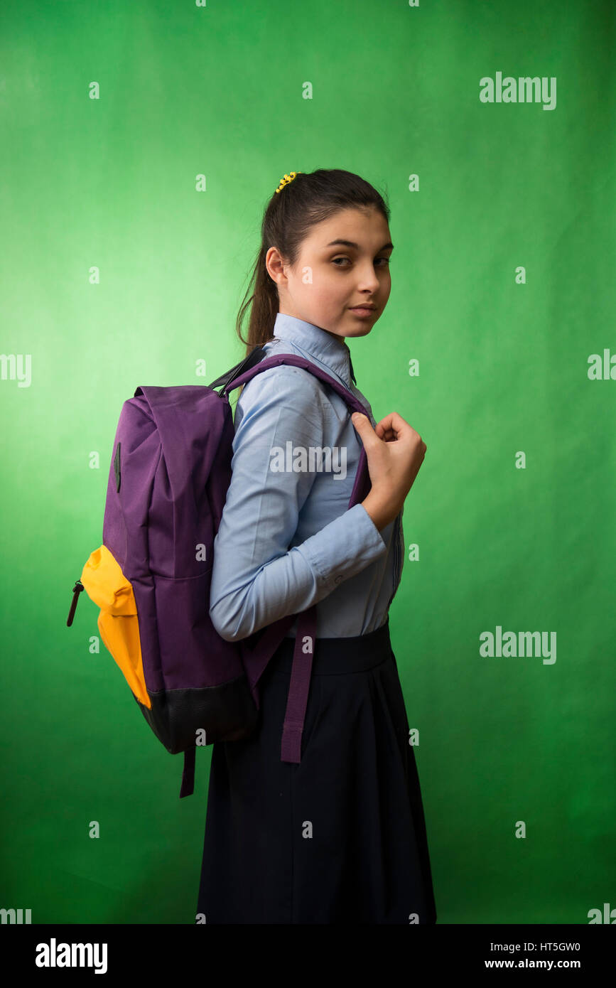 Uno teen schoolgirl con camisa azul está de pie con una mochila púrpura sobre sus hombros sobre un fondo chroma key verde Foto de stock