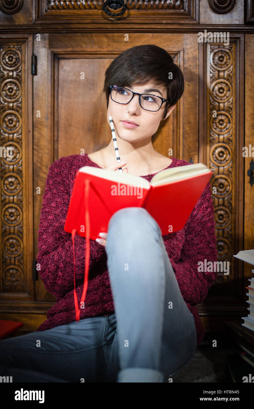Joven estudiante con expresión de cara absorbe al leer el bloc de notas rojo sentado en el piso Foto de stock
