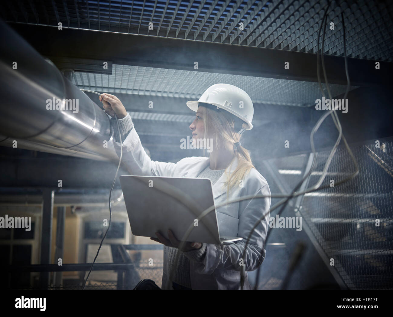 Mujer joven con casco, bata blanca de laboratorio y el portátil de pie delante de una planta industrial, Austria Foto de stock